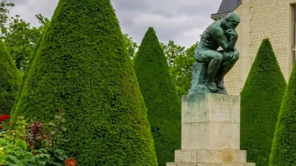 Escultura em bronze O Pensador de Auguste Rodin em um pedestal de pedra cercado por jardins franceses e arbustos esculpidos.