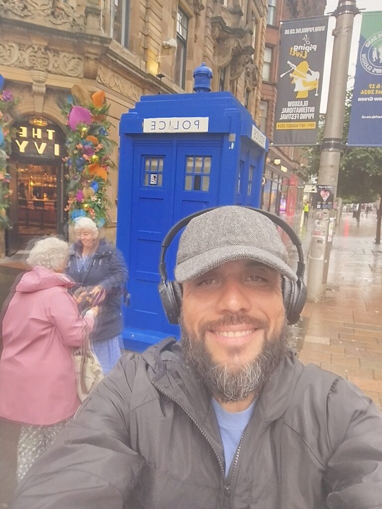 Alex Ferreira da Silva sorrindo ao lado de uma cabine de polícia azul icônica (TARDIS) em uma rua de Glasgow, Escócia.