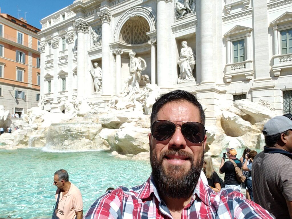 Alex Ferreira da Silva, fundador do blog Turismo Sem Fronteiras, sorrindo em frente à Fontana di Trevi em Roma, Itália.