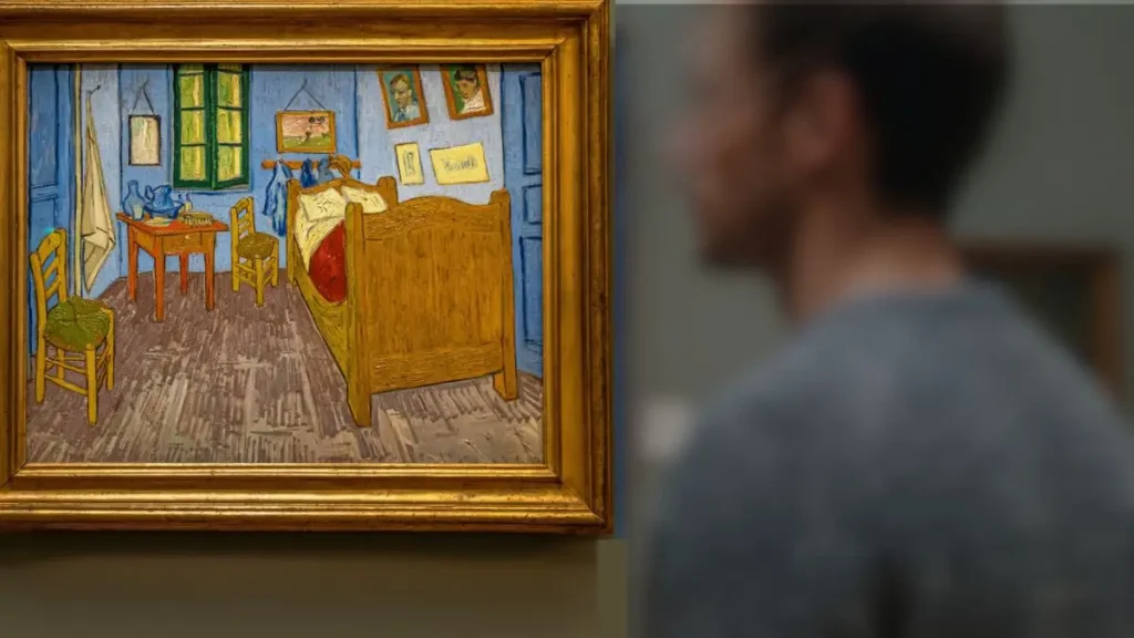 Close-up da pintura 'O Quarto de Van Gogh em Arles' com destaque para a moldura dourada e o efeito de profundidade com visitante desfocado em primeiro plano no Museu D'Orsay.