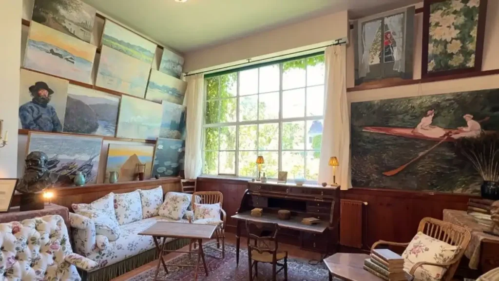 Interior do ateliê de Claude Monet na sua casa em Giverny, França. Paredes cobertas de réplicas e obras originais do pintor, com luz natural entrando por uma grande janela central, revelando a intimidade do artista para o centenário de 1926-2026.