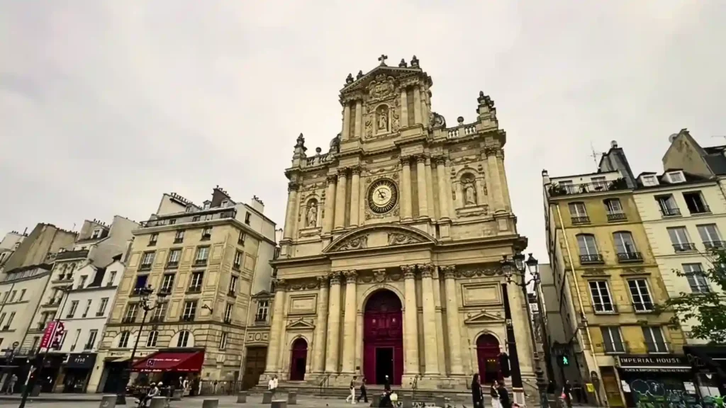 The phenomenal Baroque facade of the Église Saint-Paul-Saint-Louis church in the Marais district of Paris, showing the massive central clock, intricate religious sculptures, and towering columns, framed by nearby 'Imperial House' buildings under a soft, overcast sky.