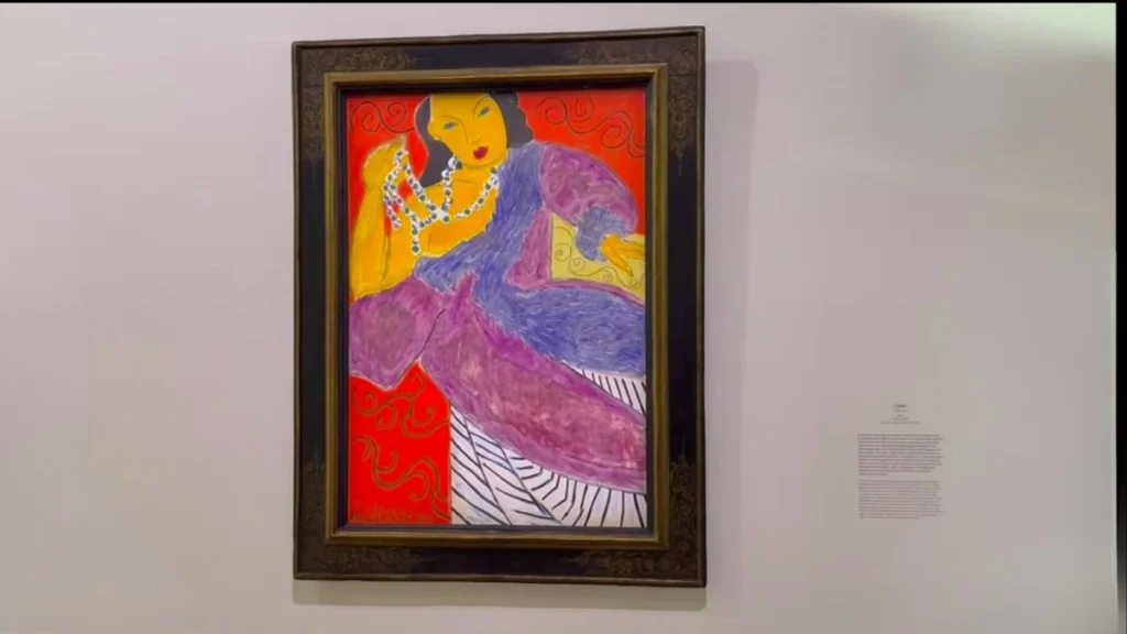 Pintura com cores vibrantes de Henri Matisse mostrando uma mulher de vestido roxo sobre um fundo vermelho e amarelo, emoldurada e exposta na parede de uma galeria de arte em Paris.